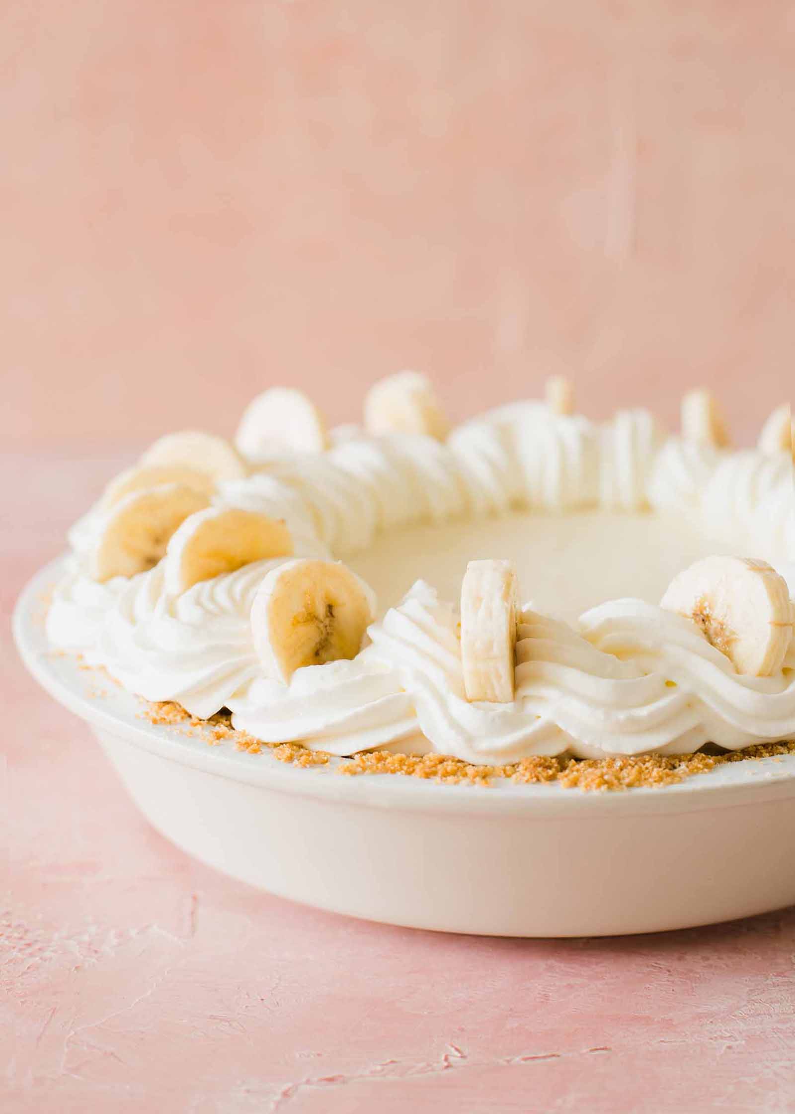 Whole banana cream pie with whipped cream swirls on top and banana slices set against a pink background.