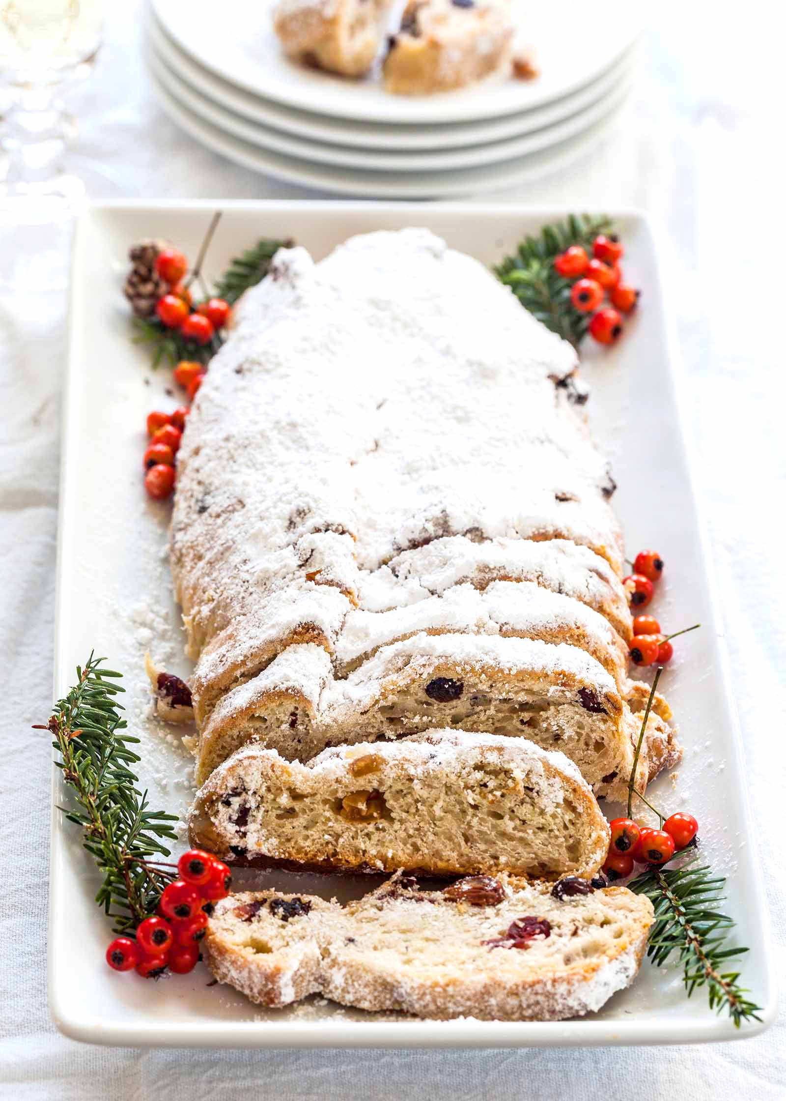 Stollen Christmas Bread filled with rum-soaked dried fruit coated with powdered sugar and sliced on a platter with a fine mesh sieve with powdered sugar with pine sprigs and berries nearby.