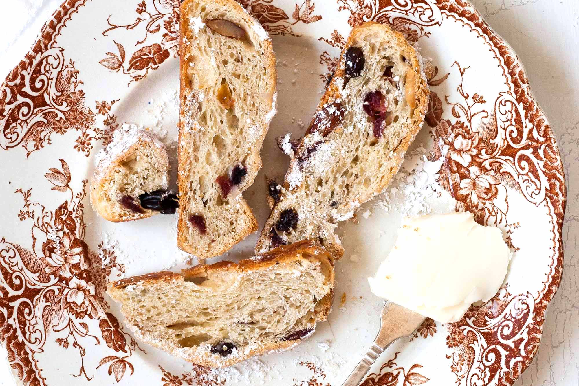 Stollen Christmas Bread filled with rum-soaked dried fruit coated with powdered sugar and sliced on a platter with a fine mesh sieve with powdered sugar in it nearby.