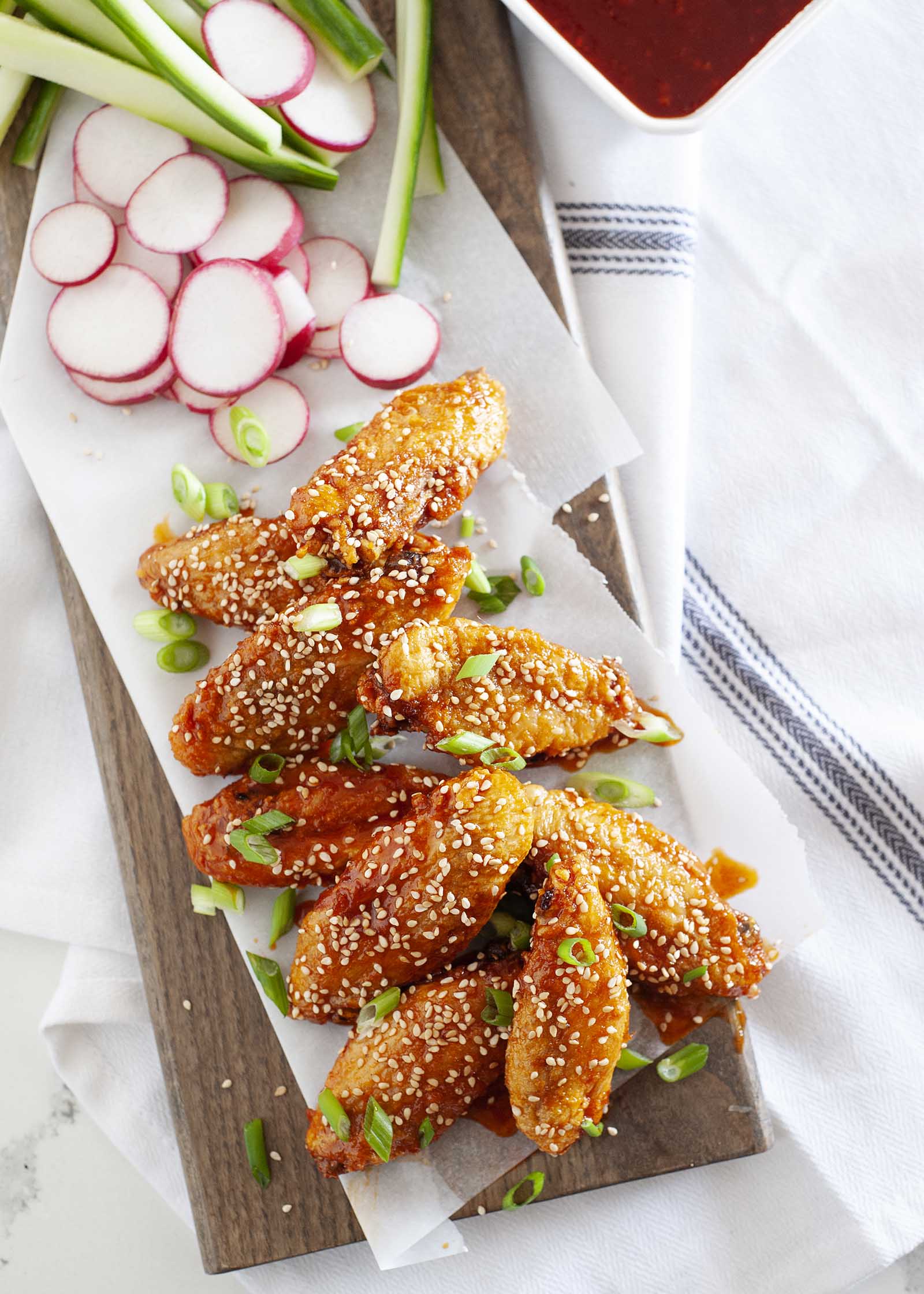 Crispy Day Air Fryer Chicken Wings with Gochujang Sauce on rectangle board sprinkled with sesame seeds with radishes, scallions and sesame dipping sauces in white dishes nearby.
