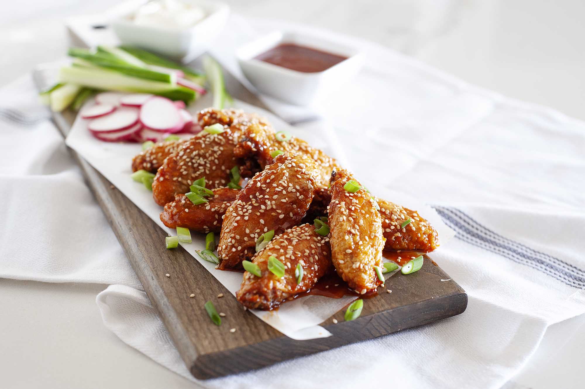Crispy Air Fryer Chicken Wings with Gochujang Sauce on rectangle board sprinkled with sesame seeds with radishes and peas nearby.