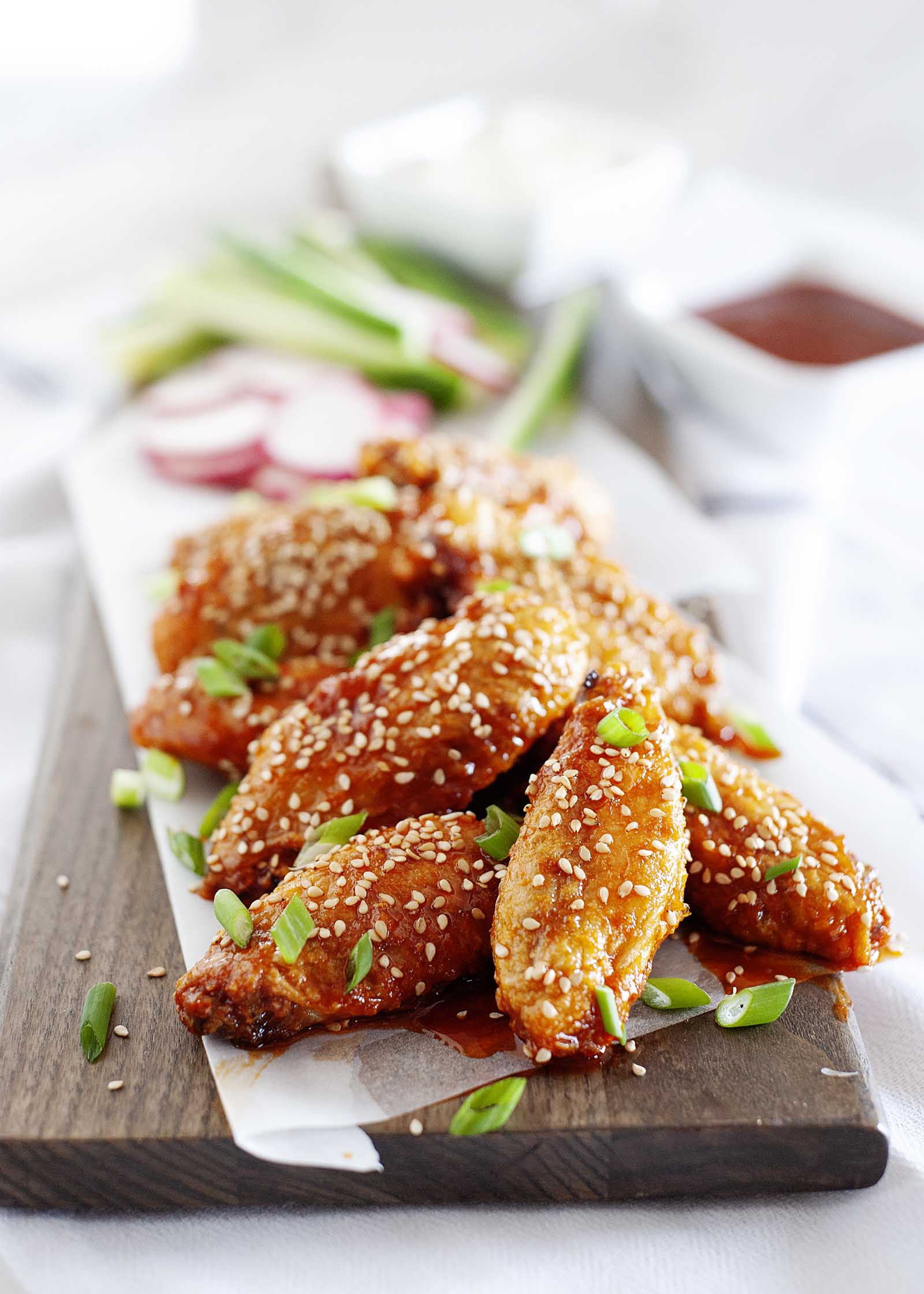 Best Air Fryer Chicken Wings with Gochujang Sauce on rectangle board sprinkled with sesame seeds with radishes, scallions and dipping sauces in white dishes nearby.