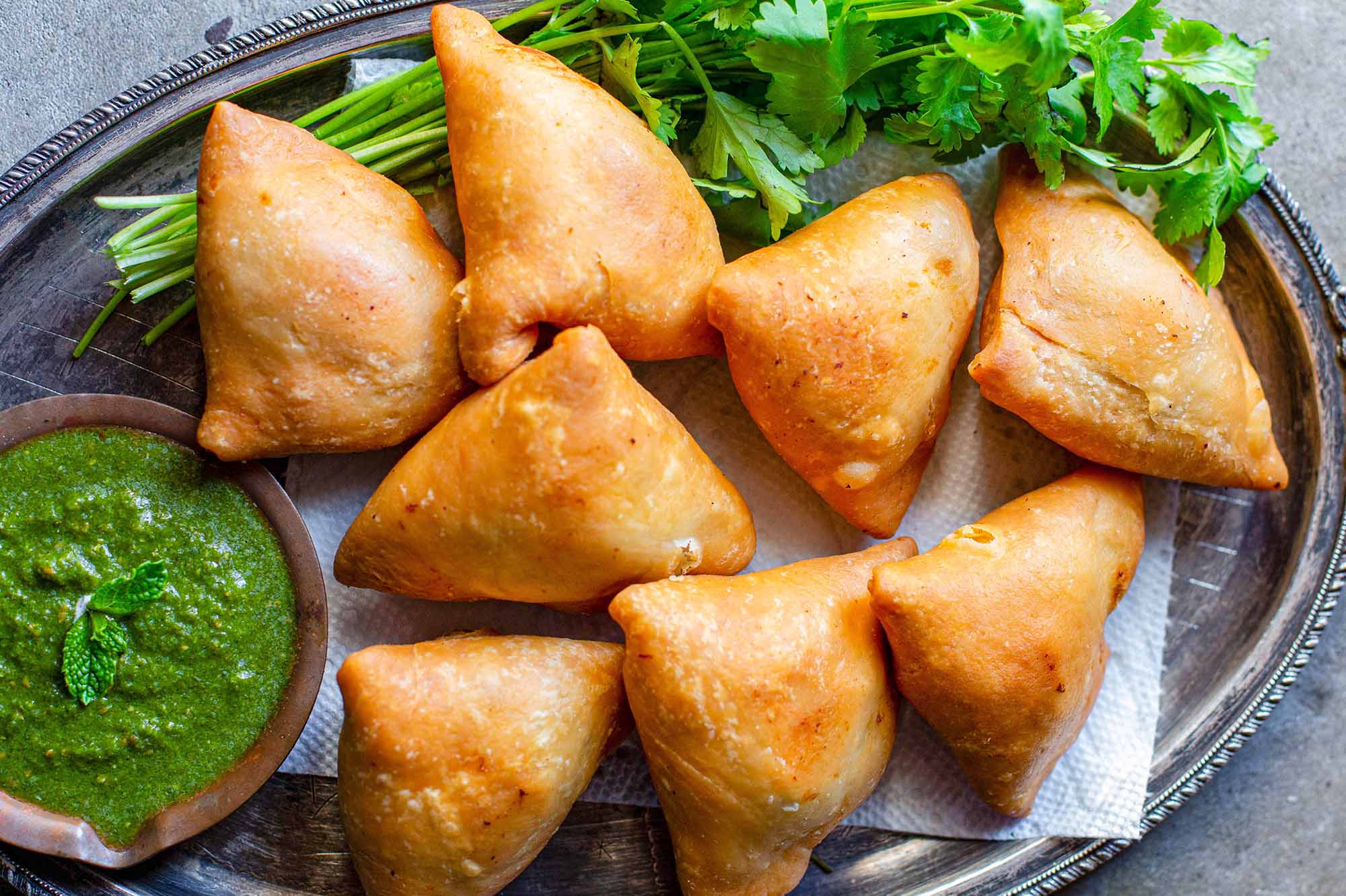 Vegetarian Samosas on a bed of herbs with one samosa filled with potato and peas alongside a cilantro mint chutney or dipping sauce in a small wooden bowl..