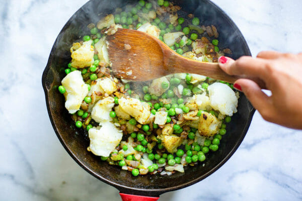 Onions, spices, peas, potatoes and jalapenos being toasted in a black skillet for vegetable samosa filling.