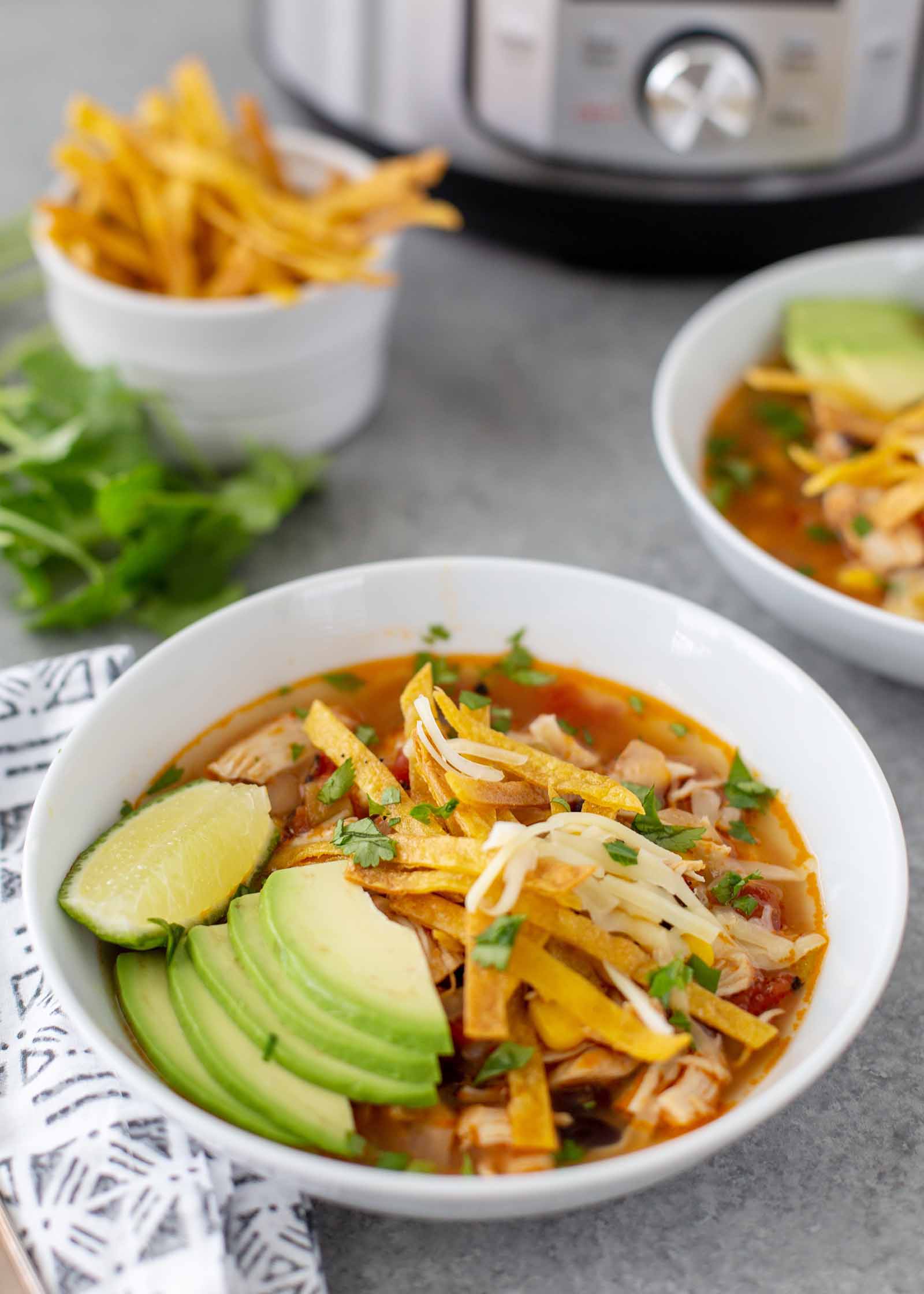 A white bowl of the best instant pot chicken tortilla soup. Each bowl has a red broth with shredded chicken, slices of fried tortilla, cheese, lime wedge and avocado slices. A couple stems of cilantro are in the bottom right corner. A ramekin has strips of fried tortillas inside and the bottom of an instant pot is visible at the top of the photo. A grey patterned linen napkin is next to the bowl. A second bowl of soup is partially visible to the right of the first bowl.