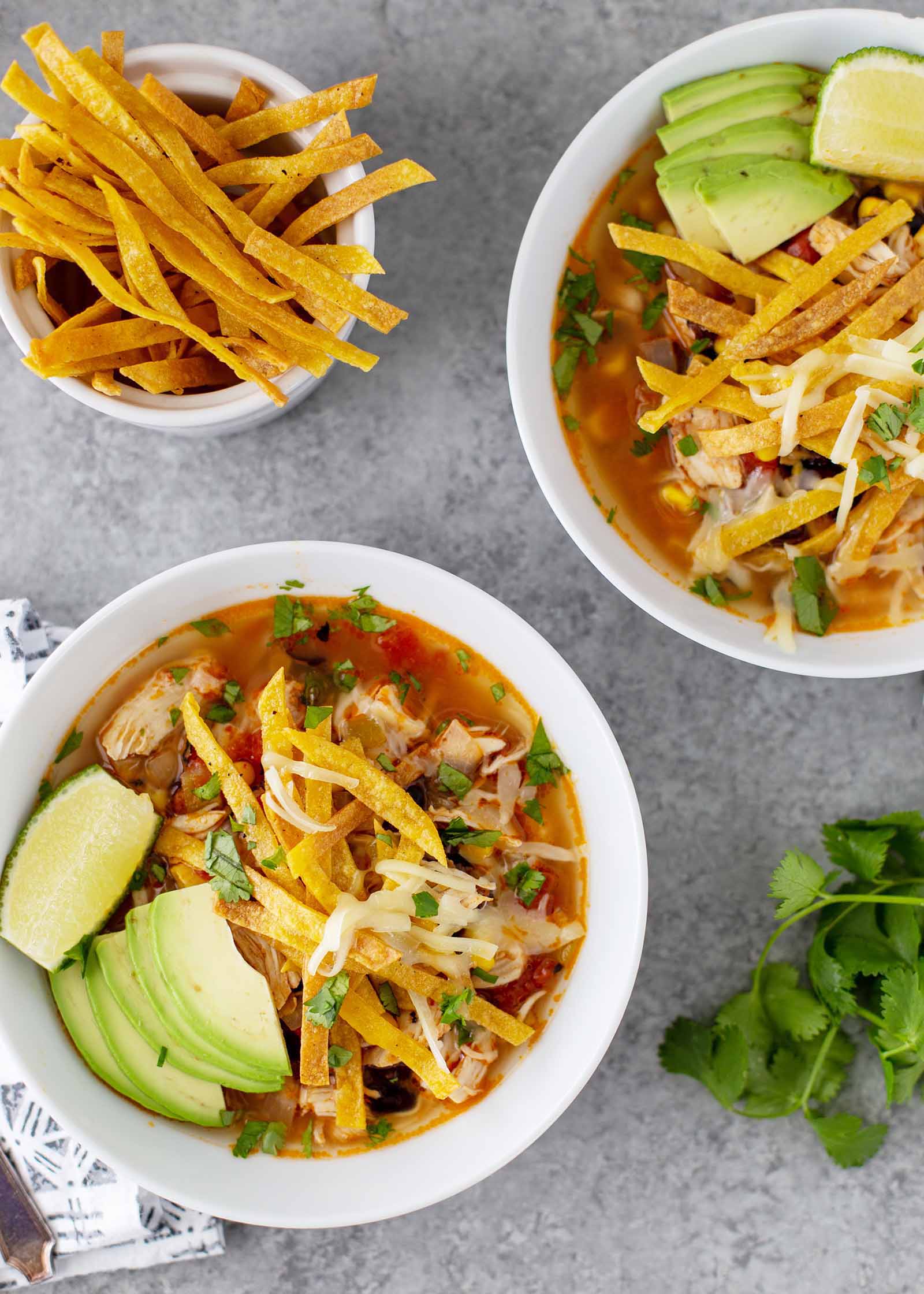 A white bowl with Instant Pot Chicken Tortilla Soup inside. A red broth with fried tortila strips, shredded chicken, chopped cilantro, shredded cheese and slices of avocado are visible in the bowl. A slice of lime is next to the avocado on the edge of the bowl. A partial view of a second bowl with the same soup inside is in the upper right corner. A white container has fried tortilla strips inside in the top left. A linen with a grey pattern is in partial view to the left of the bowl. A stem of cilantro is to the right of the bowl.