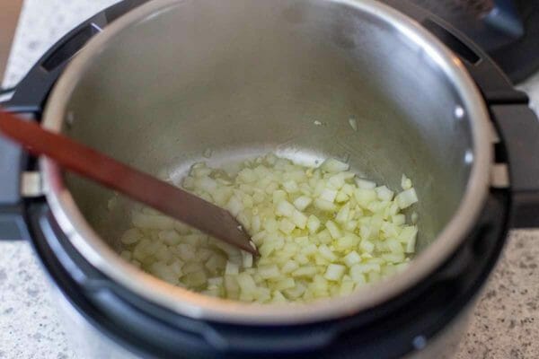 Chopped onions are being sauteed in the bottom of an instant pot along with green chilies. A wooden spoon is resting against the pot.
