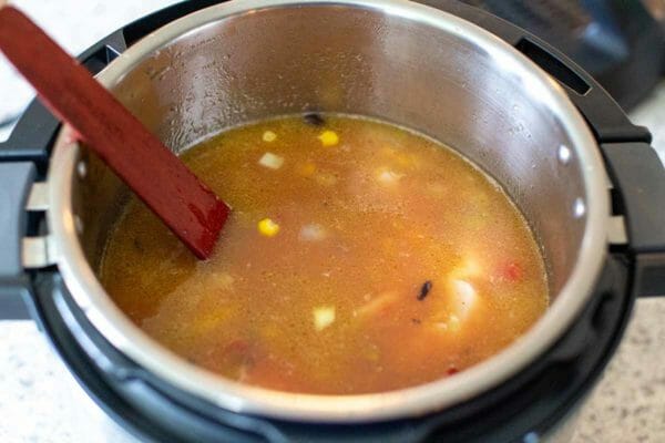 An open instant pot with pressure cooker tortilla soup inside. A golden broth with corn kernals is visible and a wooden stirring spoon is resting on the left side.