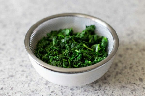 A small white bowl has a small sifter set inside with chopped frozen spinach draining.