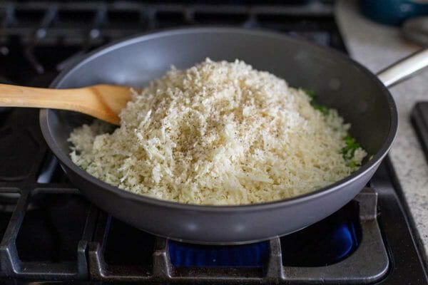 Cauliflower rice is heaped in a non-stick skillet on a gas stove. A wooden spatula is resting inside.