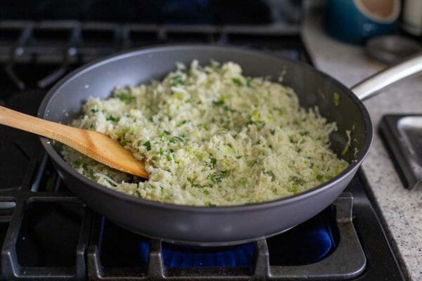 Vegan green cauliflower rice in the process of being made on in a skillet on a gas stove. Herbs are incorporated with a wooden spatula.