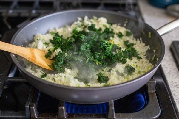 A non-stick skillet on a gas burner has cauliflower rice and chopped spinach inside. The spinach is set on top and not incorporated. A wooden spatula rests inside the skillet.