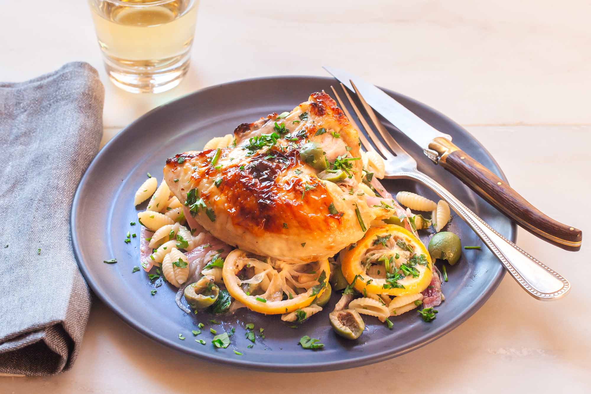 Yogurt marinated lemon chicken with pasta, olives and herbs on a rimmed plate. A fork and knife rest on the right edge of the slate plate. A grey linen is folded to the left of the plate an a tumbler of white wine is above the plate to the left.