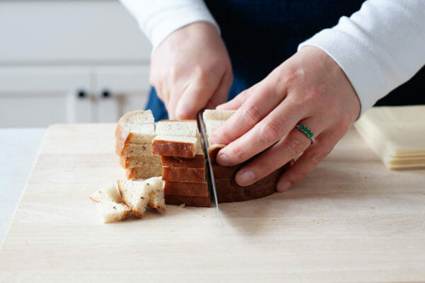 A person with a blue apron and long sleeved white shirt is holding a chef's knife and cubing rye bread on a wooden cutting board.