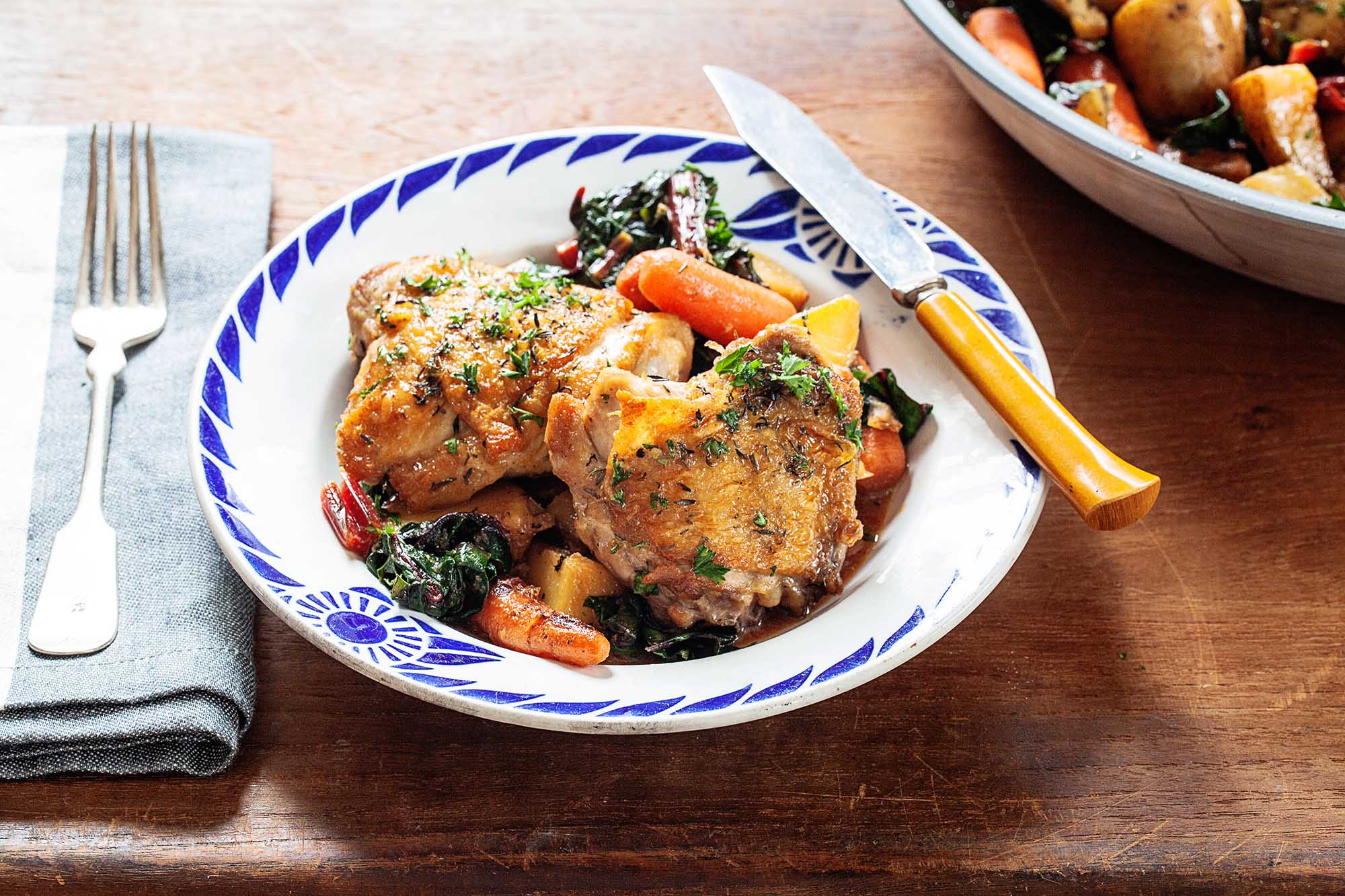 Two crispy skillet chicken thighs with baby carrots and wilted greens. The shallow bowl has a blue pattern around the edge. A yellow handled knife is on the right edge. A folded napkin has a fork on top. More of the skillet chicken thighs with potatoes, carrots and greens in a bowl partially visible in the upper right corner.