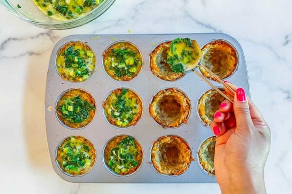 Filling hash brown egg bites with egg spinach filling. A hand is using a tablespoon to fill the crusts in the muffin tin.