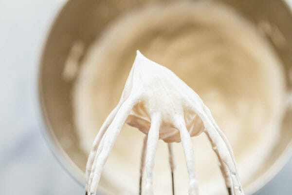 Close up of the top of a whisk with a stiff peak of whipped cream. A metal bowl with whipped cream is out of focus in the background.