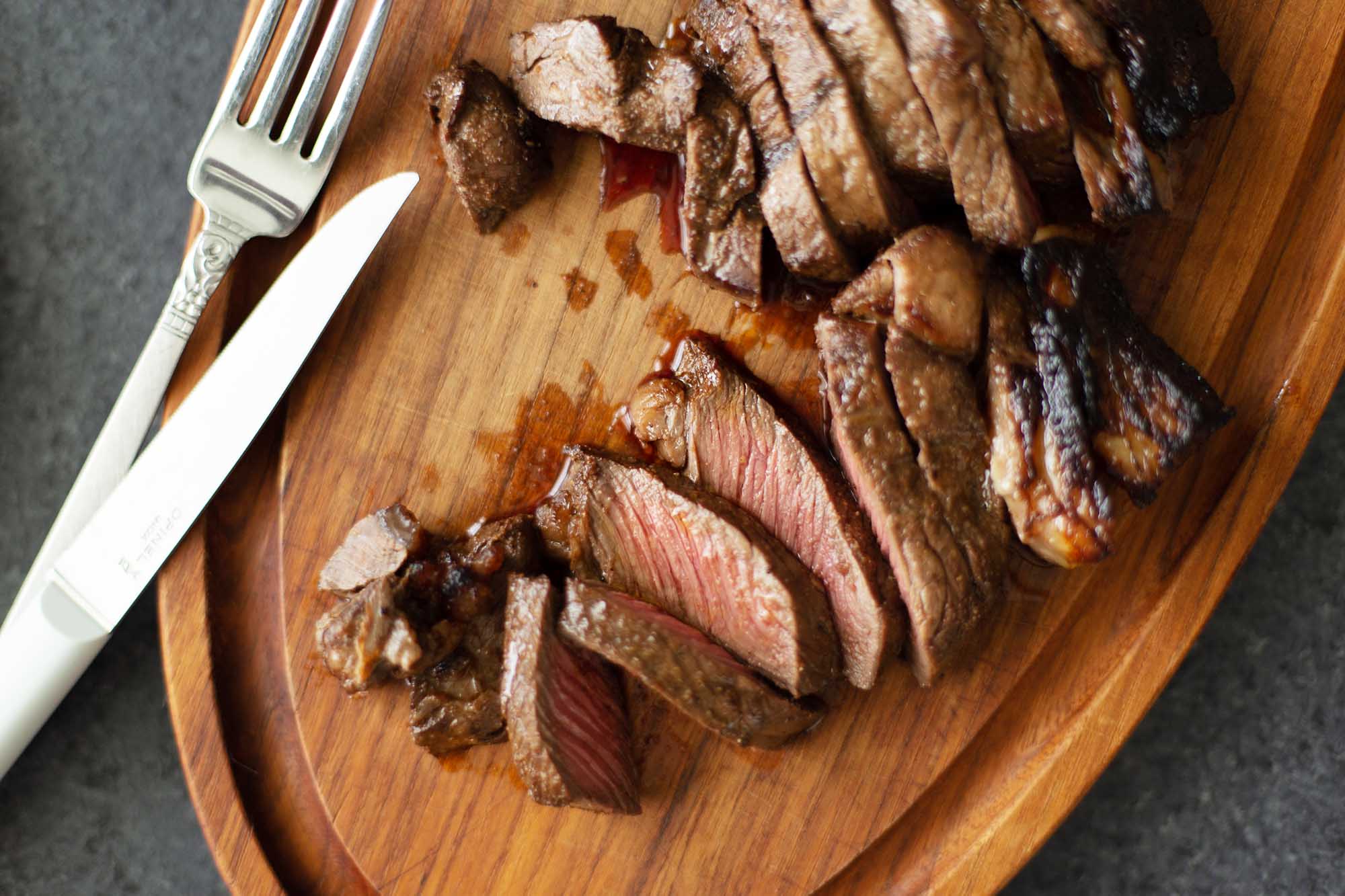 Overhead view of sliced oven broiled steak and silverware on a wooden cutting board.