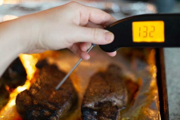 A hand is holding a thermapen that reads "132" and inserted into an oven broiled steak on a baking sheet in the oven.