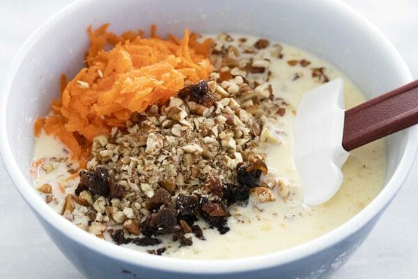The best carrot cake pancake batter is being mixed together with a flexible spatula. The smooth batter is getting shredded carrots, chopped pecans and raisins in the bowl.