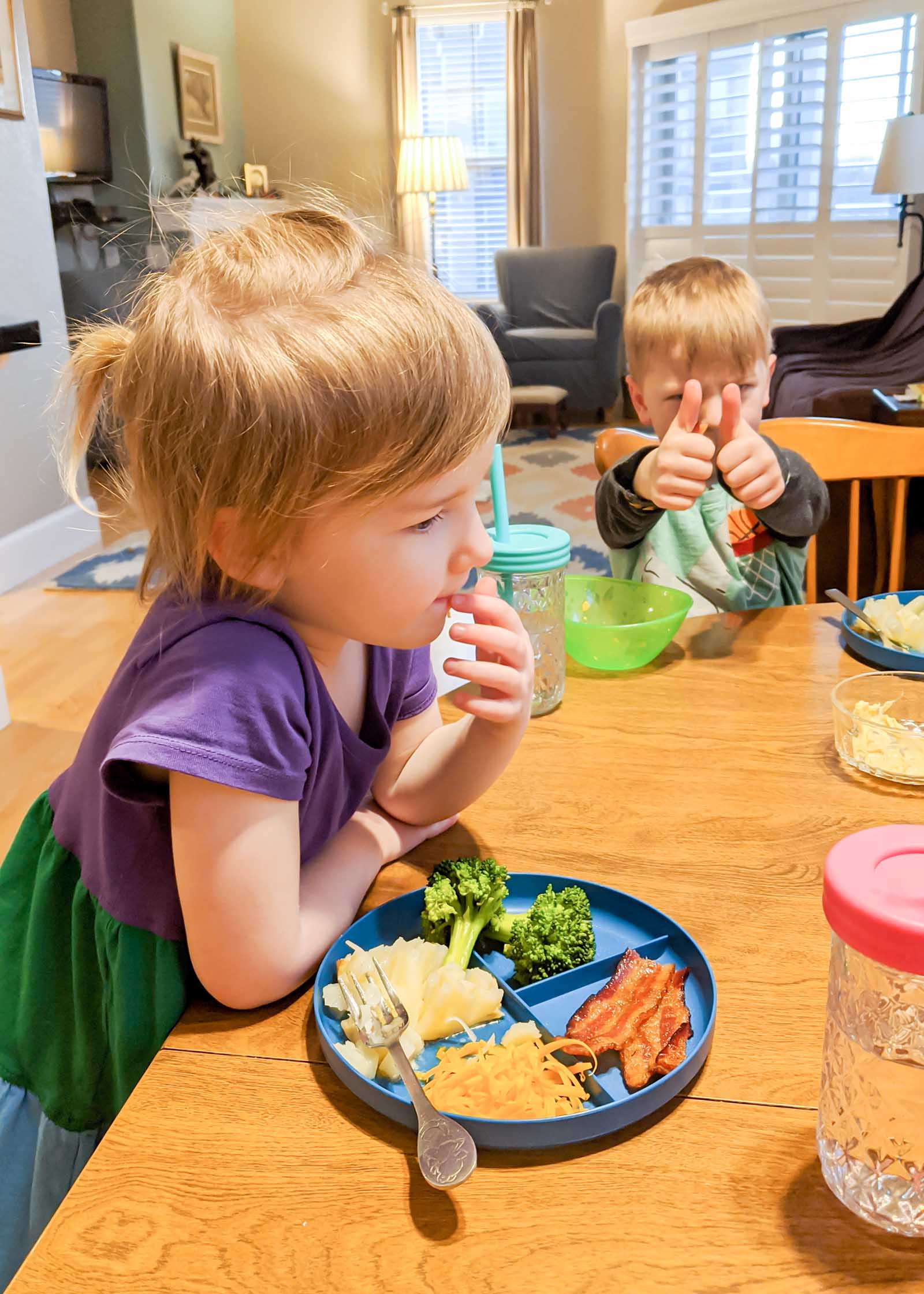 A young girl and boy are sitting at a kitchen table with plates and drinks in front of them. One plate is split into four sections. Each section has one type of food on it. Bacon, shredded cheddar cheese, mashed potato and cooked broccoli is on the plate. The boy is giving a thumbs up sign at his chair.