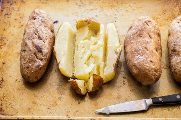 Four baked russet potatoes are on a worn baking sheet. The skin is salted and a paring knife is laid underneath the potatoes. One potato is cut down each side and across the top revealing the creamy potato inside.