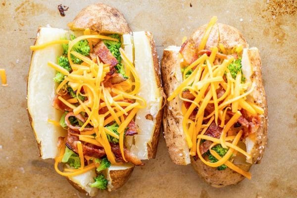 Two loaded baked potatoes with bacon on a worn baking sheet. The russet potatoes are split open with broccoli, chopped bacon and unmelted, shredded cheddar cheese.