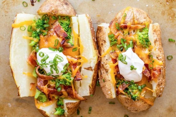 Two quick and easy loaded baked potatoes on a baking sheet. The russet potatoes are split down the middle and loaded with bacon, cheddar cheese, sliced green onions, broccoli and a dollup of sour cream in the center of the potato.