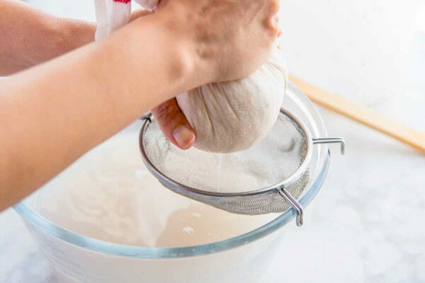 Rice in a cheesecloth is being squeezed over a small sift into a glass bowl to show how to make horchata.