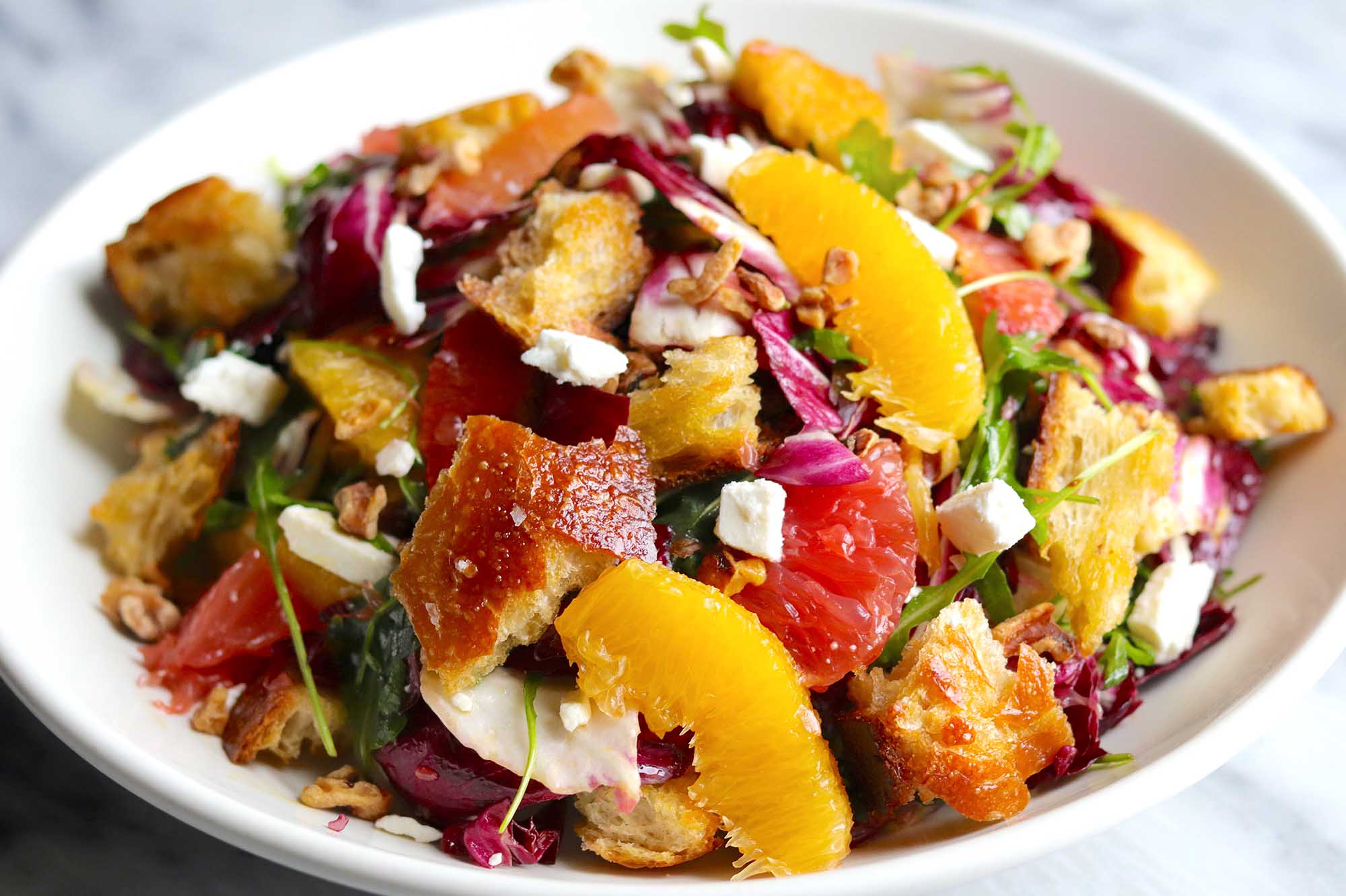 Side view of a winter salad in a white, wide bowl. Supremed orange and grapefruit, radicchio, croutons and feta cheese crumbles are visible.