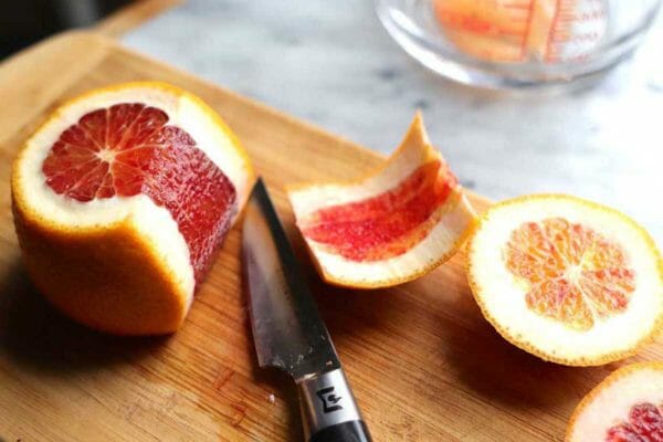 A side view of a blood orange with the top sliced off and the pith and peel partially cut off of it. The top and side peel is to the right of the knife.