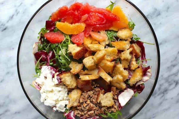 An overhead view of the inside of a large glass bowl. Elements of the bitter green and citrus salad are inside. Homemade croutons are in the middle of the bowl and sit on top of the arugula and radicchio. Feta is in the lower left and supremed oranges and grapefruit are above the croutons.