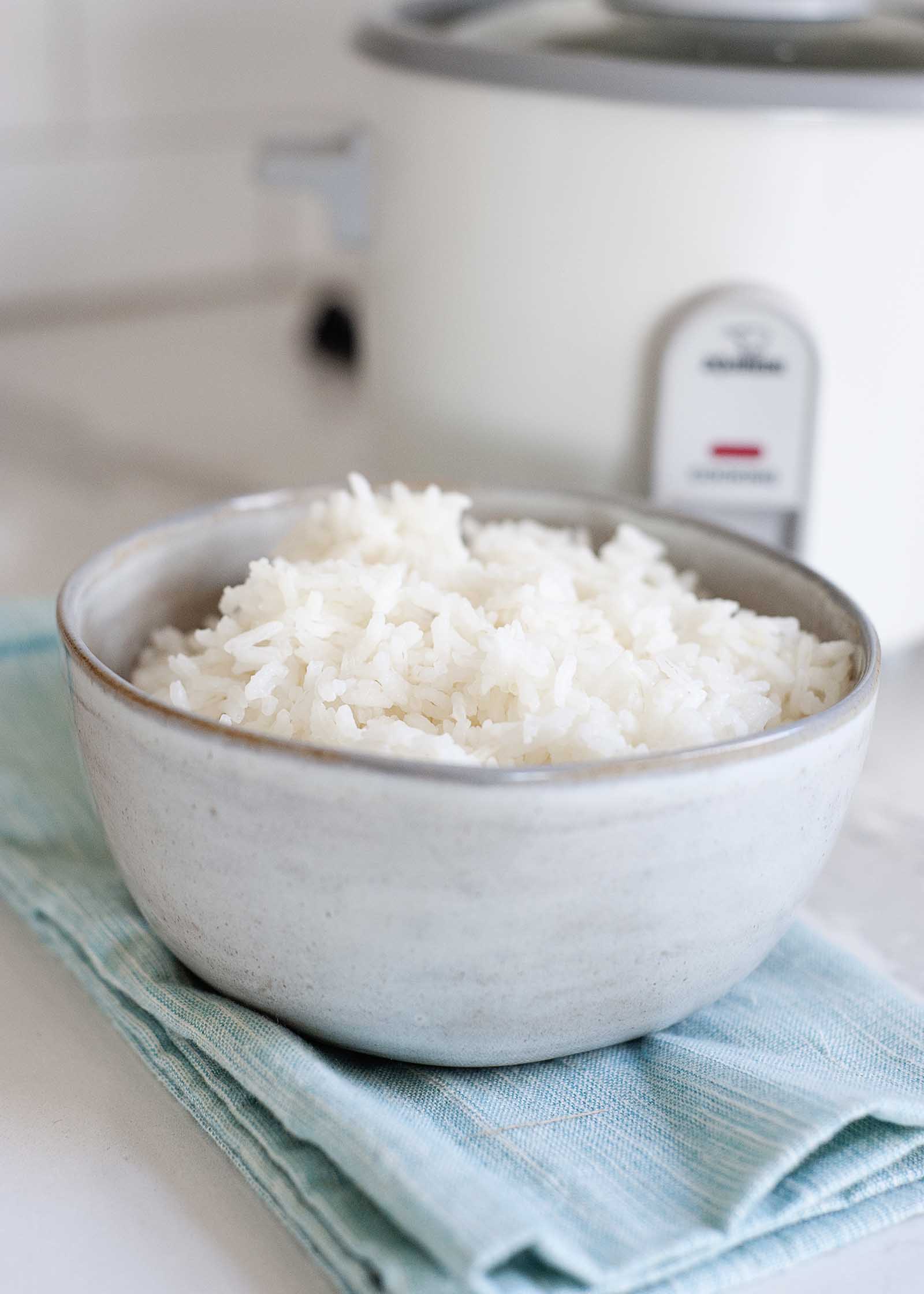 Vertical image of a ceramic bowl filled with white rice on a white counter. A teal dish towel is under the bowl of rice and a white rice cooker is behind the bowl of rice.