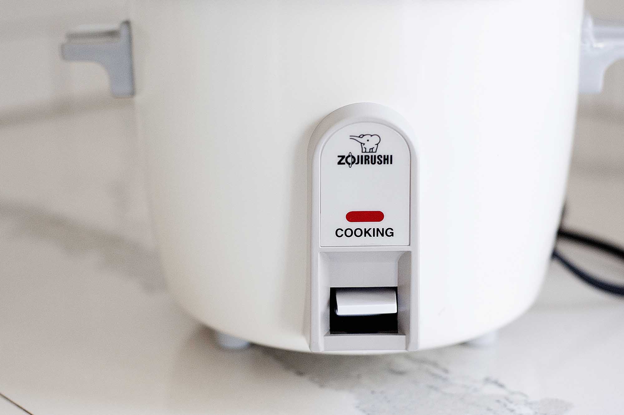 Horizontal image of a side view of the bottom half of a Zojirushi rice cooker on a white countertop.