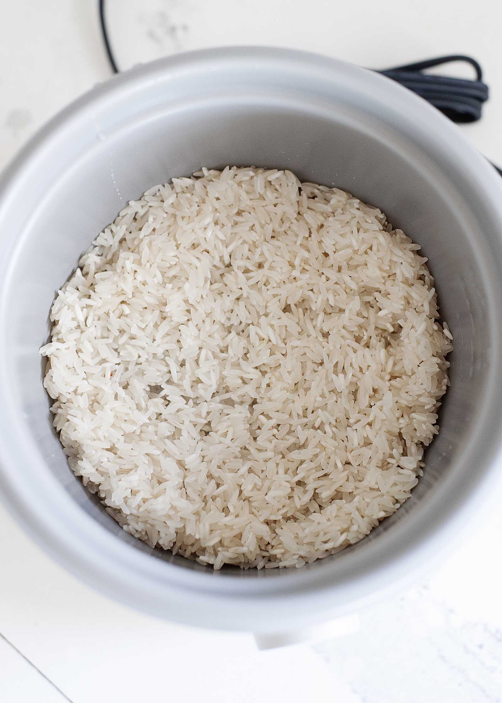 Overhead view of the inside of a white rice cooker with uncooked rice inside. The cord is in partial view behind the cooker.