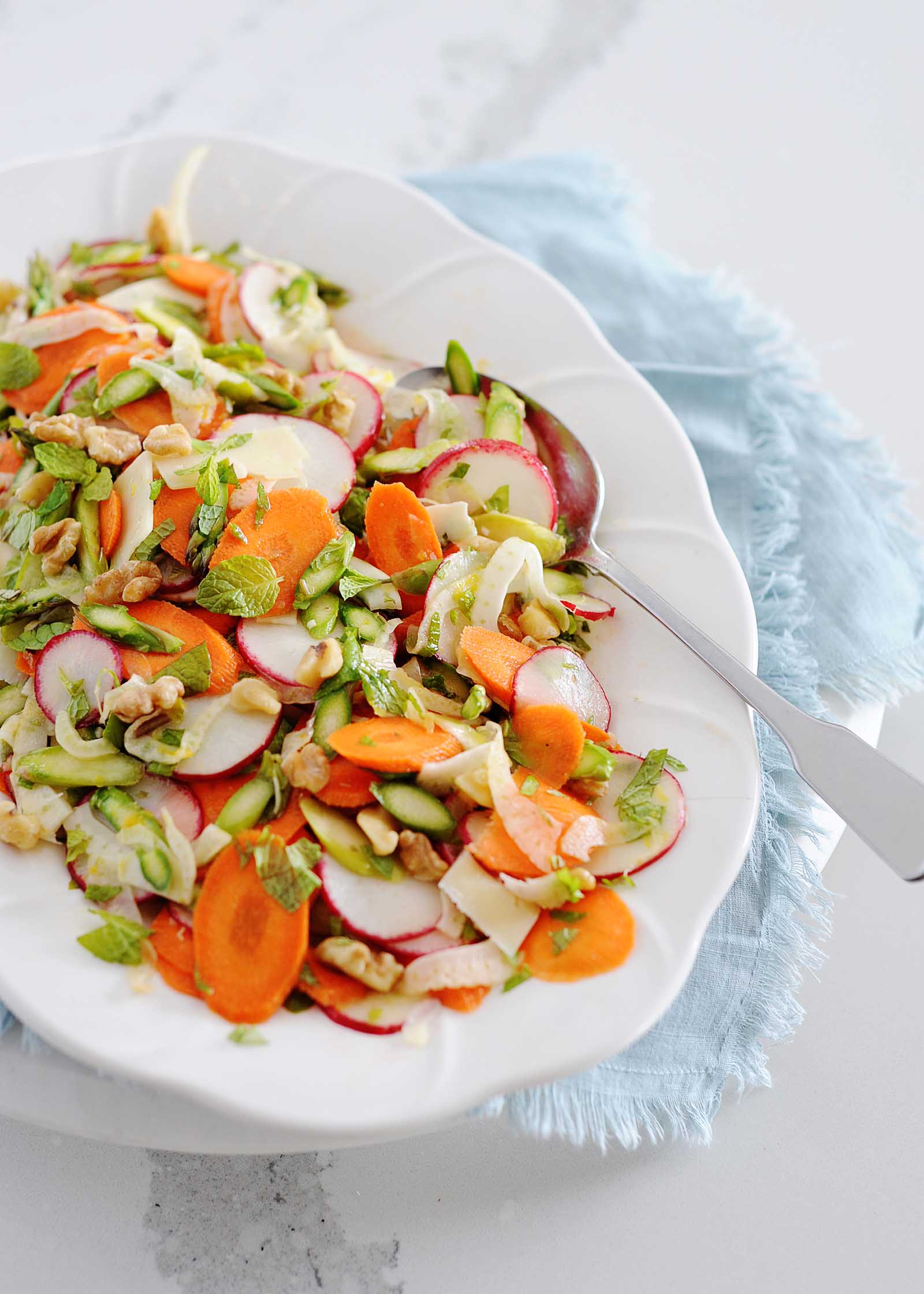Overhead angled view of a white platter with a shaved vegetable salad of thinly sliced carrot, radish, fennel and asparagus mixed together. Mint leaves are scattered around the top. The platter is on a light blue linen on a white counter and a silver serving spoon is on the right side of the platter.