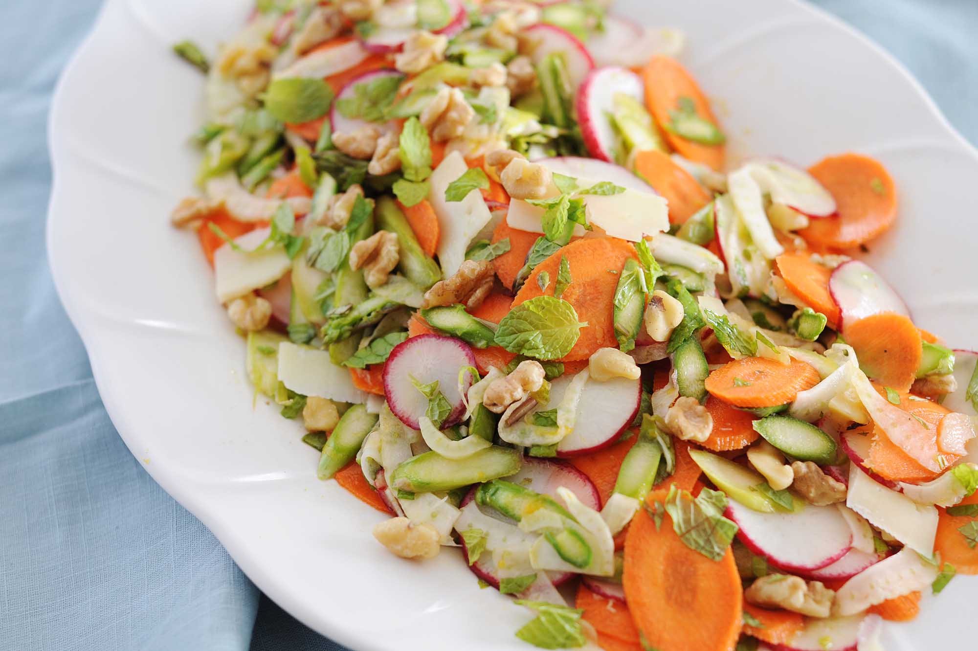 A white platter with an easy spring salad. Thinly sliced carrot, radish, fennel and asparagus are mixed together and mint leaves and chopped nuts are scattered around the top. The platter is on a light blue linen.