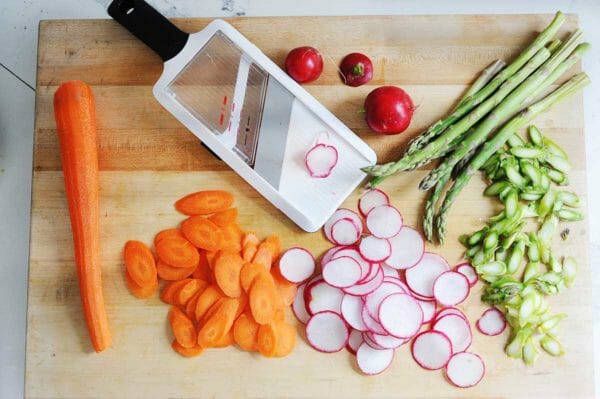 Wooden cutting board with ingredients for spring salad with shaved vegetables. A mandalin has a slice of radish on it and a pile of sliced radishes, carrots and asparagus nearby. Whole peeled carrot is on the left of the board. Whole radishes and asparagus stems are on the right of the mandalin.