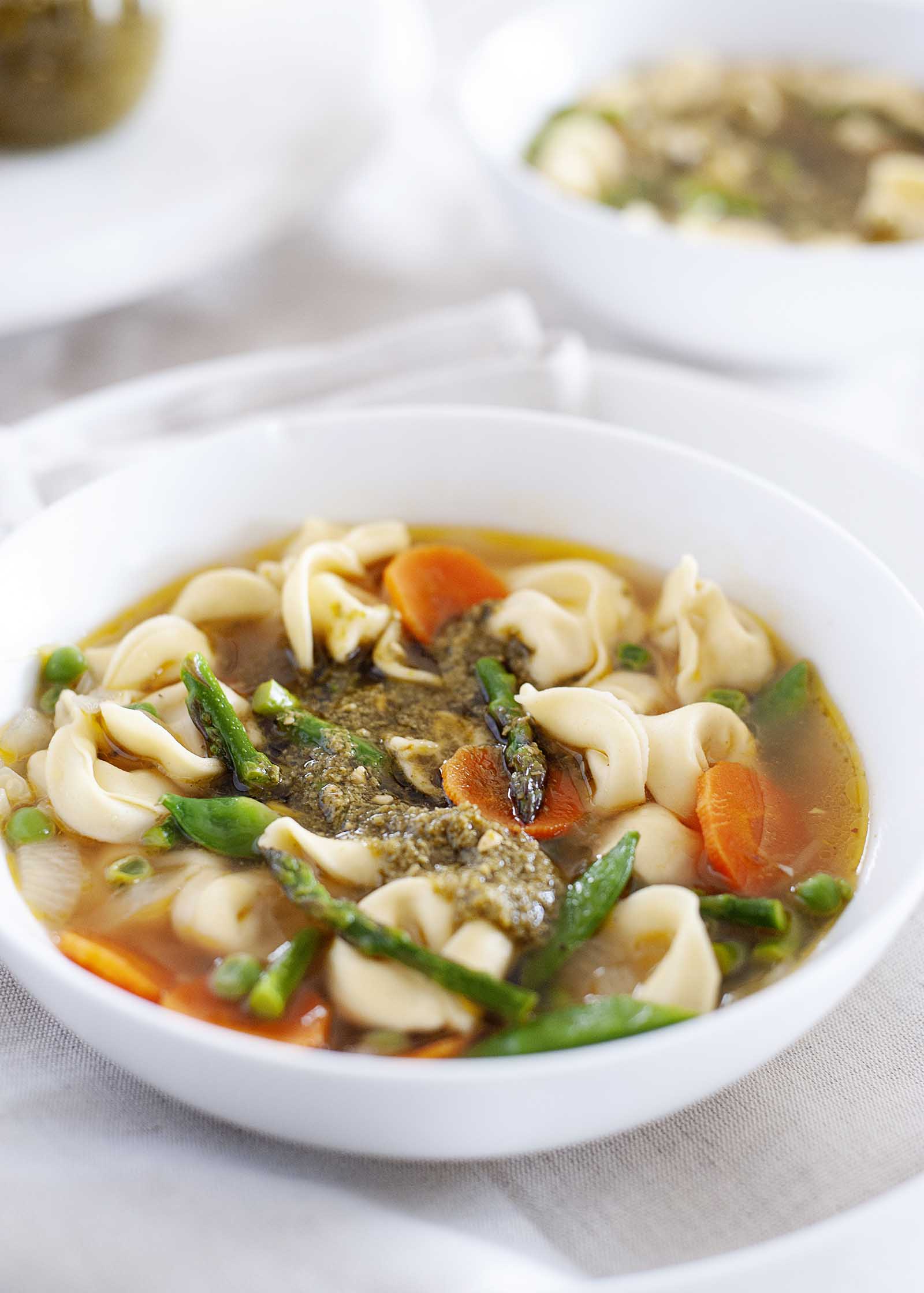 Bowl of vegetarian soup with tortellini in a white bowl on a table with a second bowl in the background. Chopped asparagus, sliced carrots and pesto are visible with the tortellini.