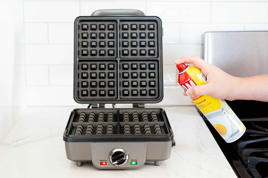Cooking spray is being sprayed on an open waffle maker.
