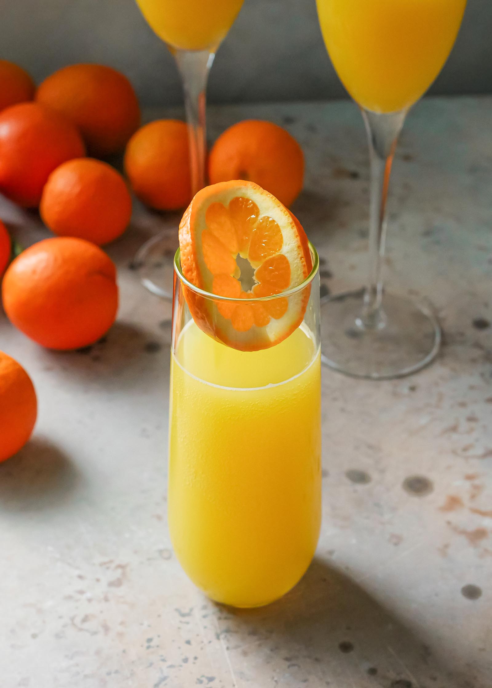Eeasy mimosa in a glass with an orange slice inside. Two flutes of mimosa are behind the glass and oranges are behind the drinks.