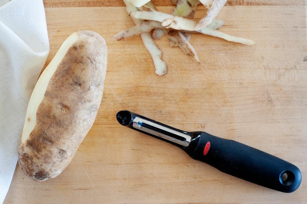 A russett potato with some of the skin peeled off and a black vegetable peeler to the left.