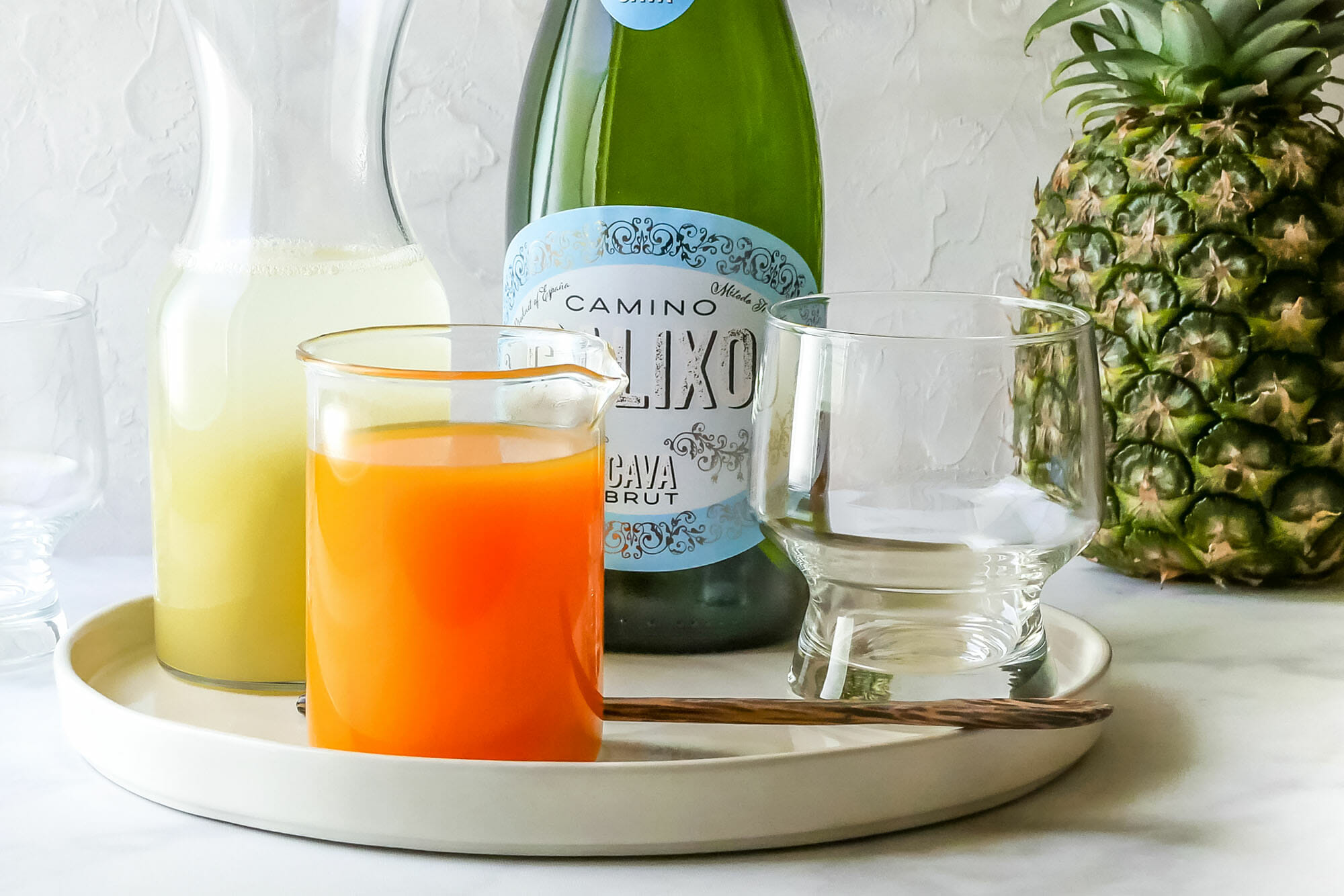 A white tray has pineapple and mango juice, a champagne glass, and bottle of sparkling wine. A whole pineapple is behind the tray.