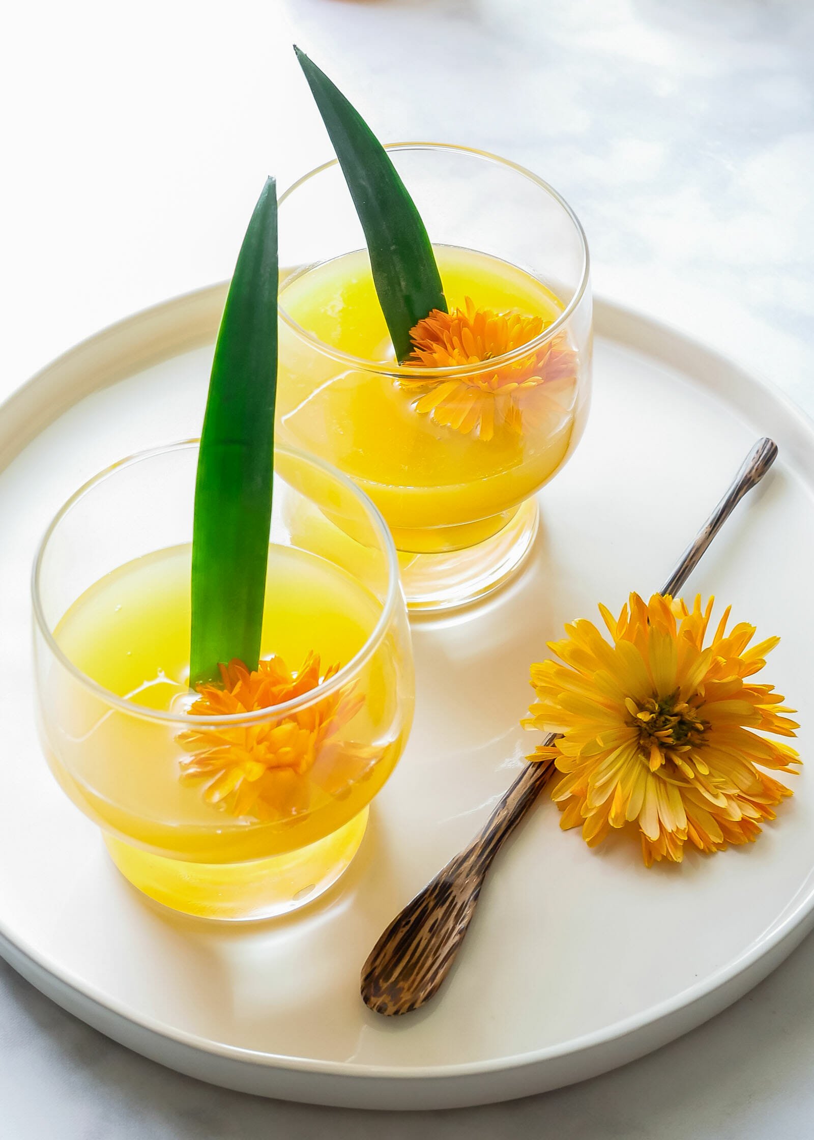 A white tray has two glasses of cava mimosa with an orange flower an d green pineapple leaf decorating each one. A stir stick and a third flower is on the tray.