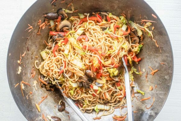 A steel wok with the best vegetable lo mein stir fried together using a pair of tongs.