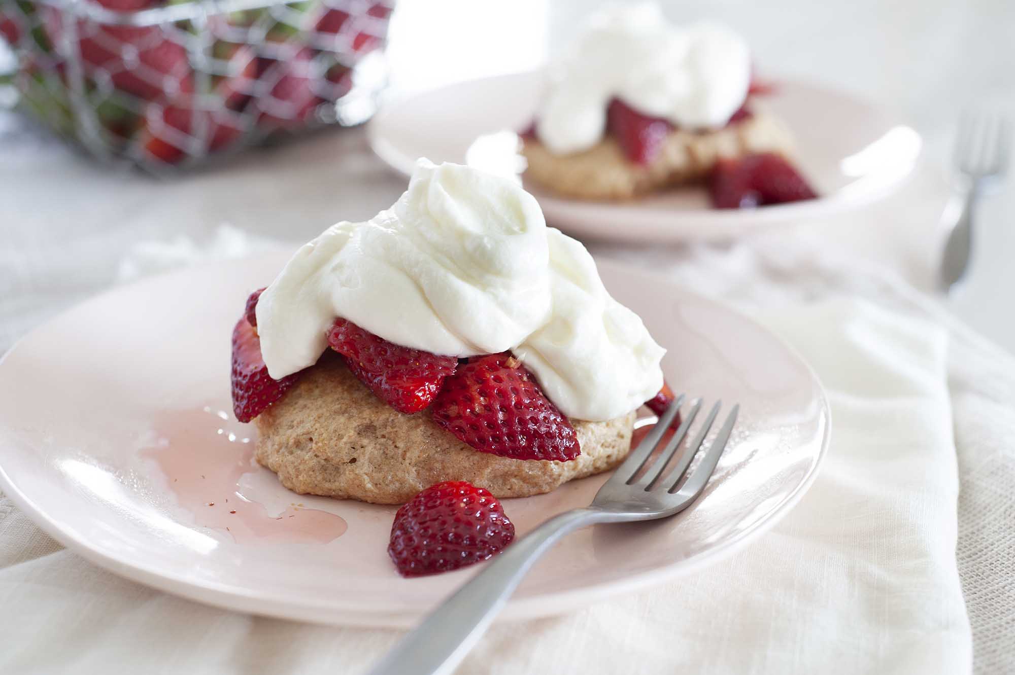 Two plates with healthier strawberry shortcake set on a table with white linens. Shortcake is piled with sliced strawberries and whipped yogurt topping. A wire basket of strawberries is to the left of the plates.