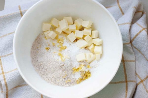 A white bowl with cubed butter, flour and lemon zest to make light strawberry shortcake. The bowl is on a yellow striped linen.