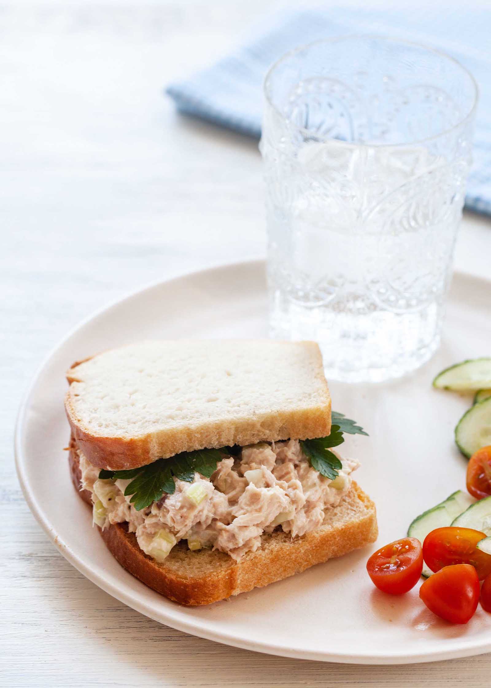 White bread with quick and easy tuna salad inside. A glass of water, sliced cucumbers and halved tomatoes are also on the plate.