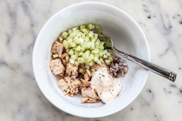 White bowl with chopped celery, mayo, tuna and red onion along with a fork.