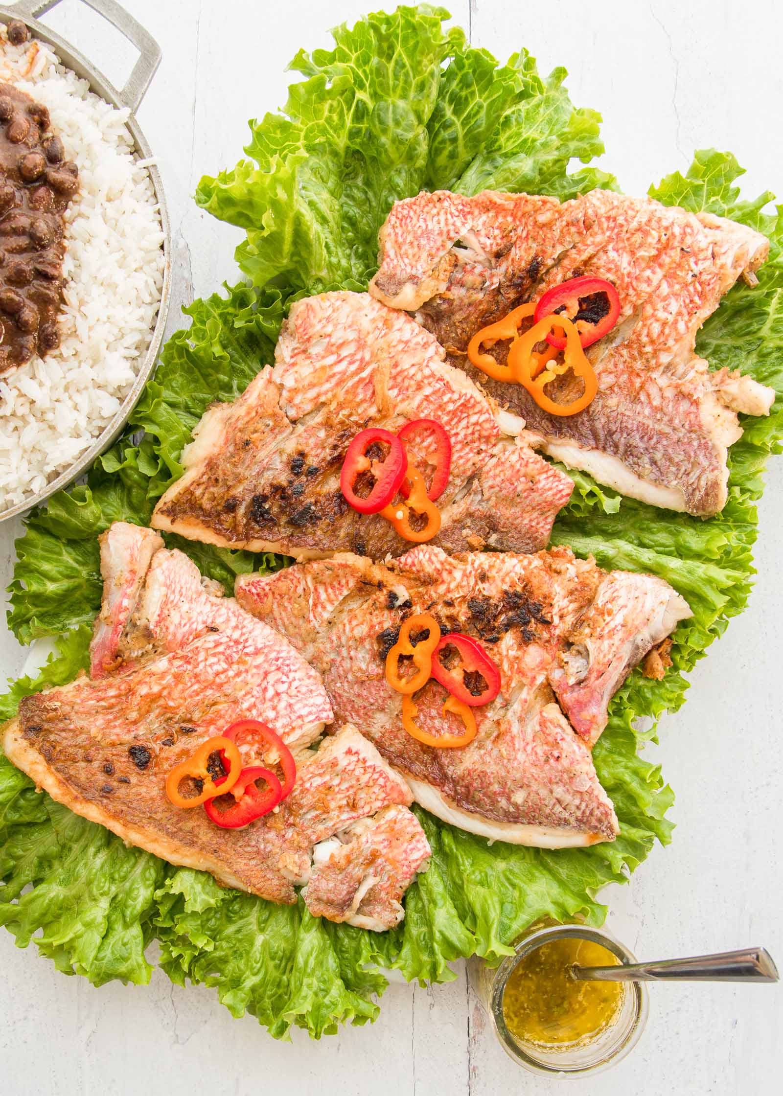 A platter of red snapper with garlic sauce is sitting vertically on a white table. Four crispy skinned snapper fillets are on top of a bed of frilly lettuce. Sliced red and orange peppers are on each fillet. A large pot of rice and black beans is in partial view to the left. A small jar of garlic sauce is in the bottom right corner.